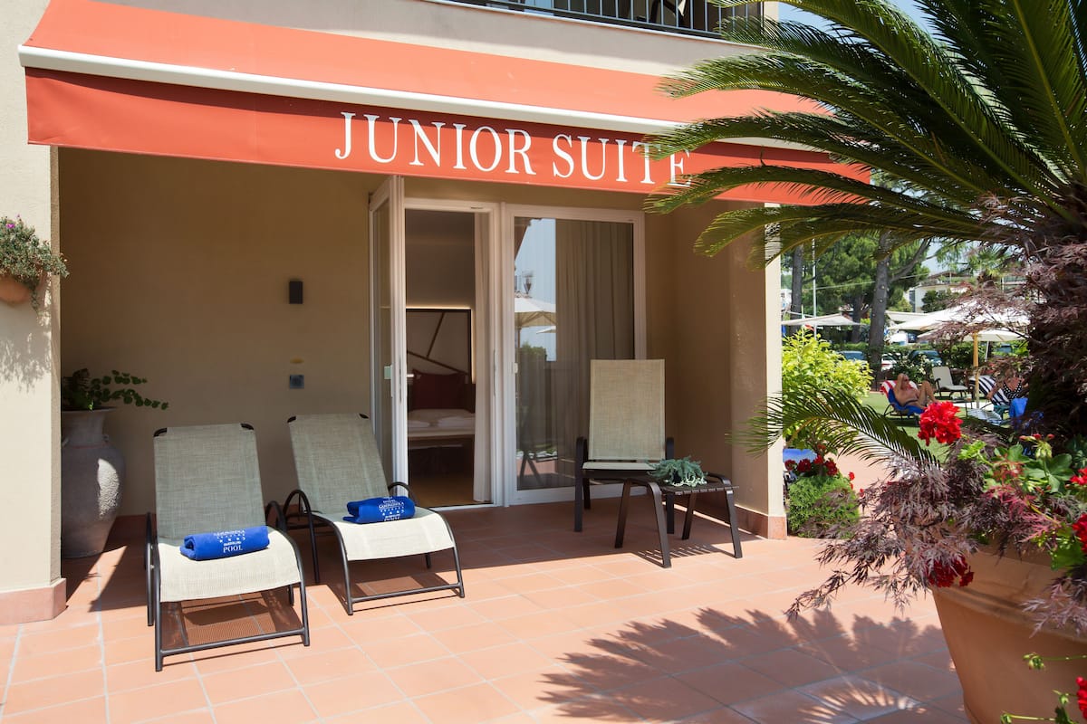 A hotel entrance with a sign reading "JUNIOR SUITE". There are two lounge chairs with blue towels, a potted plant, and a palm tree.