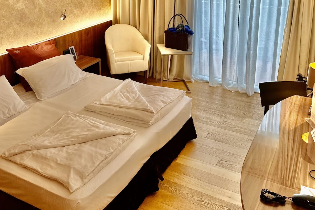 A hotel room with a large bed, a desk, a chair, a small table, and a window with curtains.