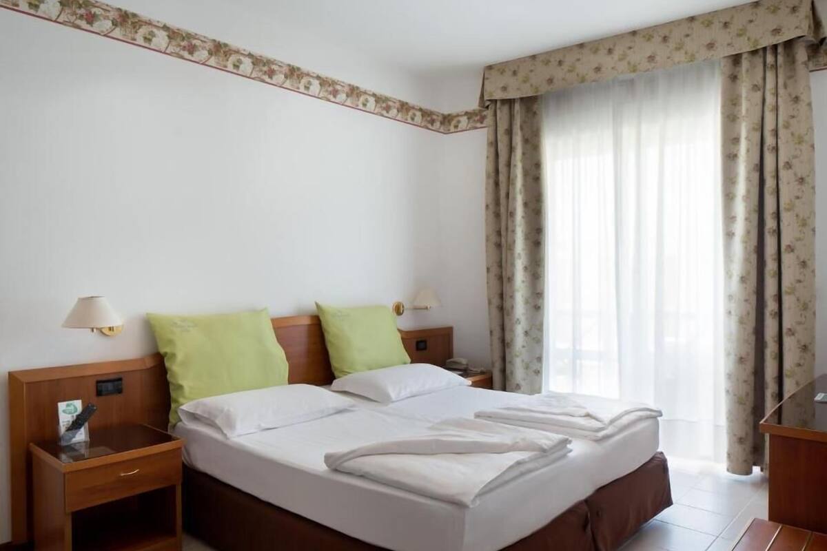 A hotel room with a large bed, two bedside tables, a nightstand, a lamp, and a view of the outdoors through a window with curtains.