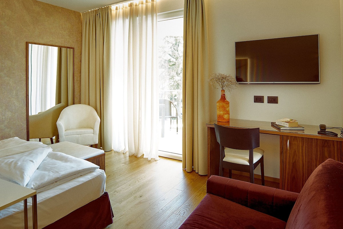 A hotel room with a bed, two chairs, a desk, a TV, and a window with curtains.