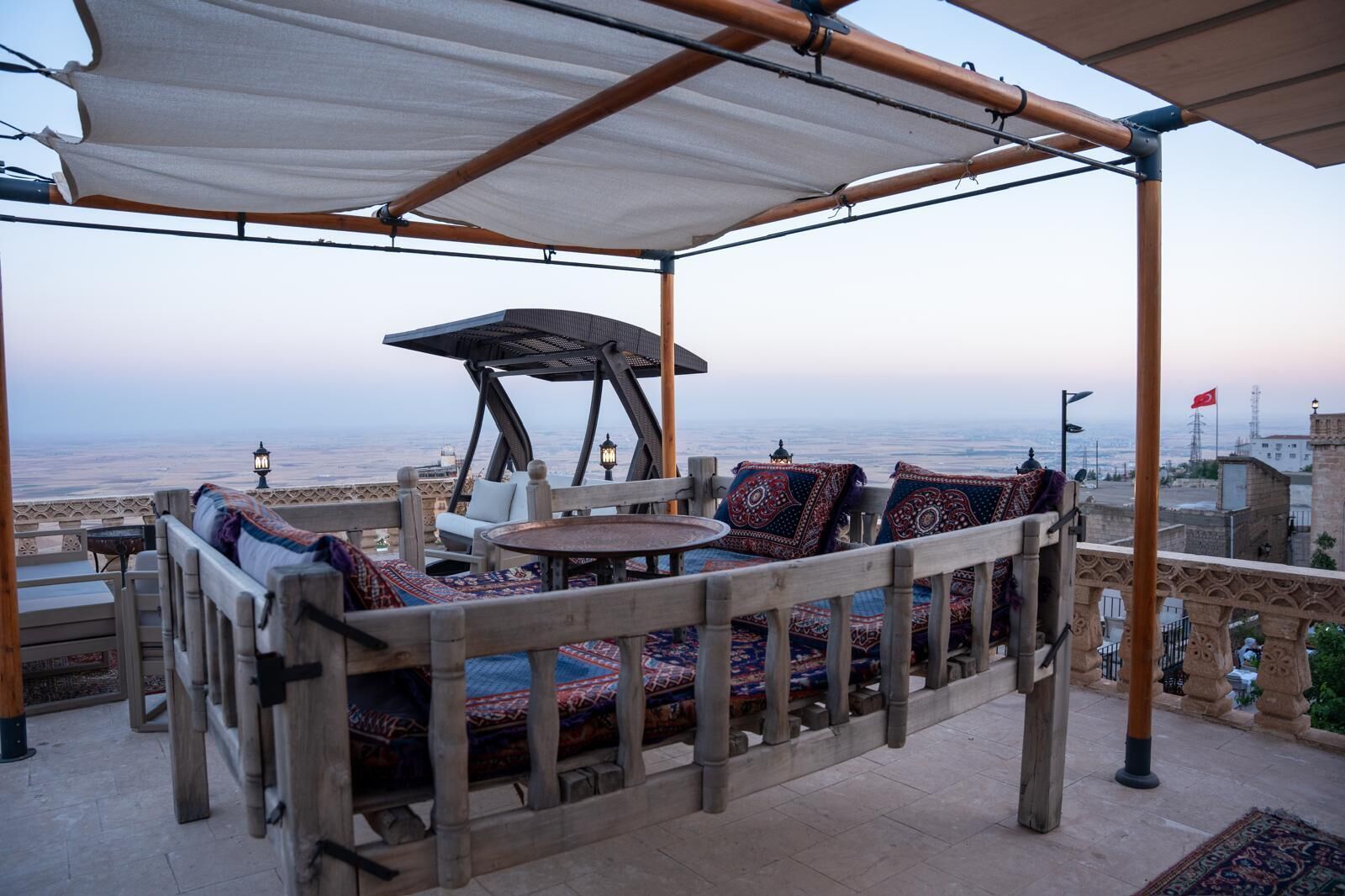 An outdoor seating area with a view of the cityscape, featuring a wooden pergola, cushioned seating, and a small table.