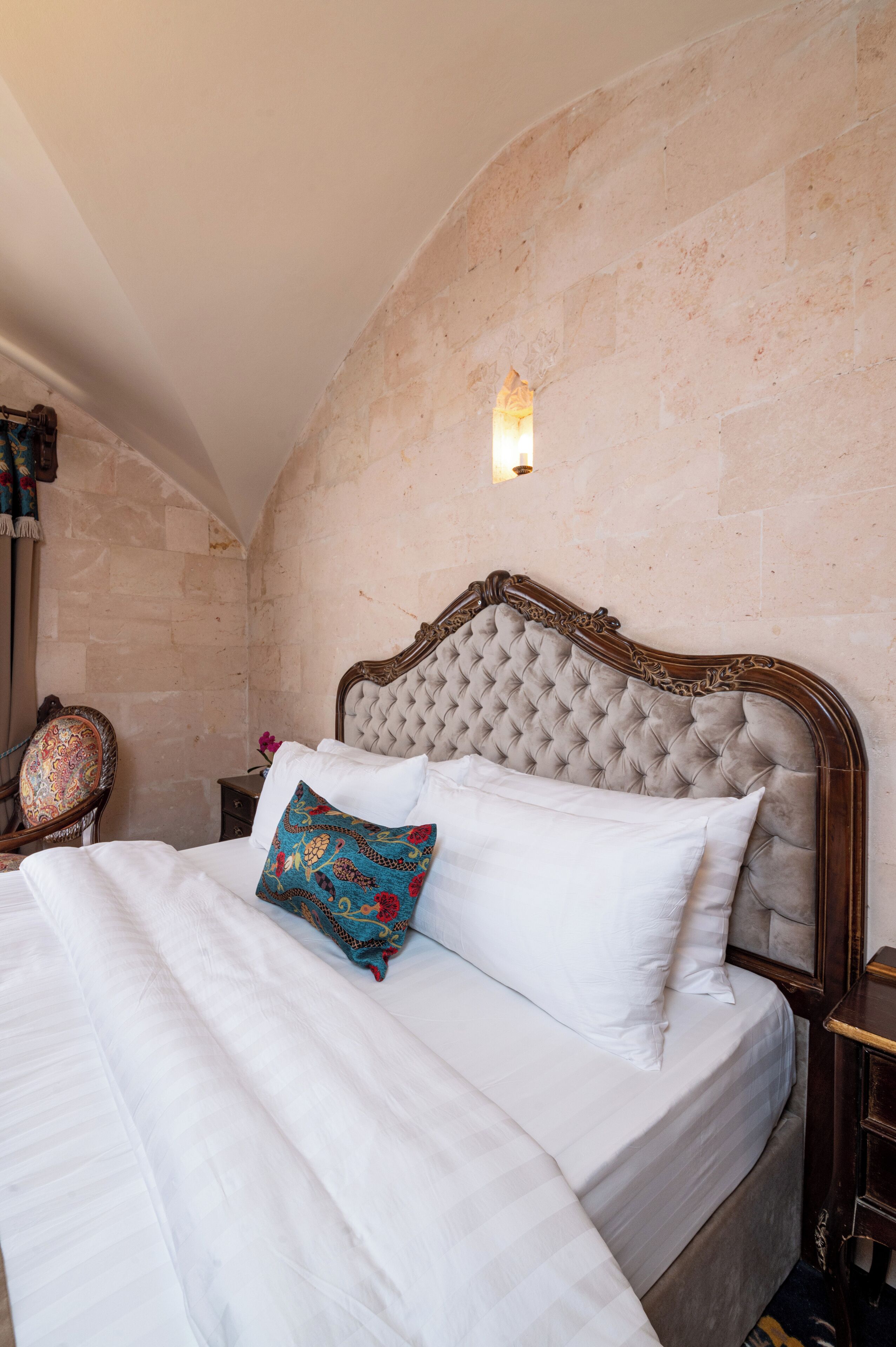 A neatly made bed with white linens and a decorative pillow, set against a stone wall with a wooden headboard.