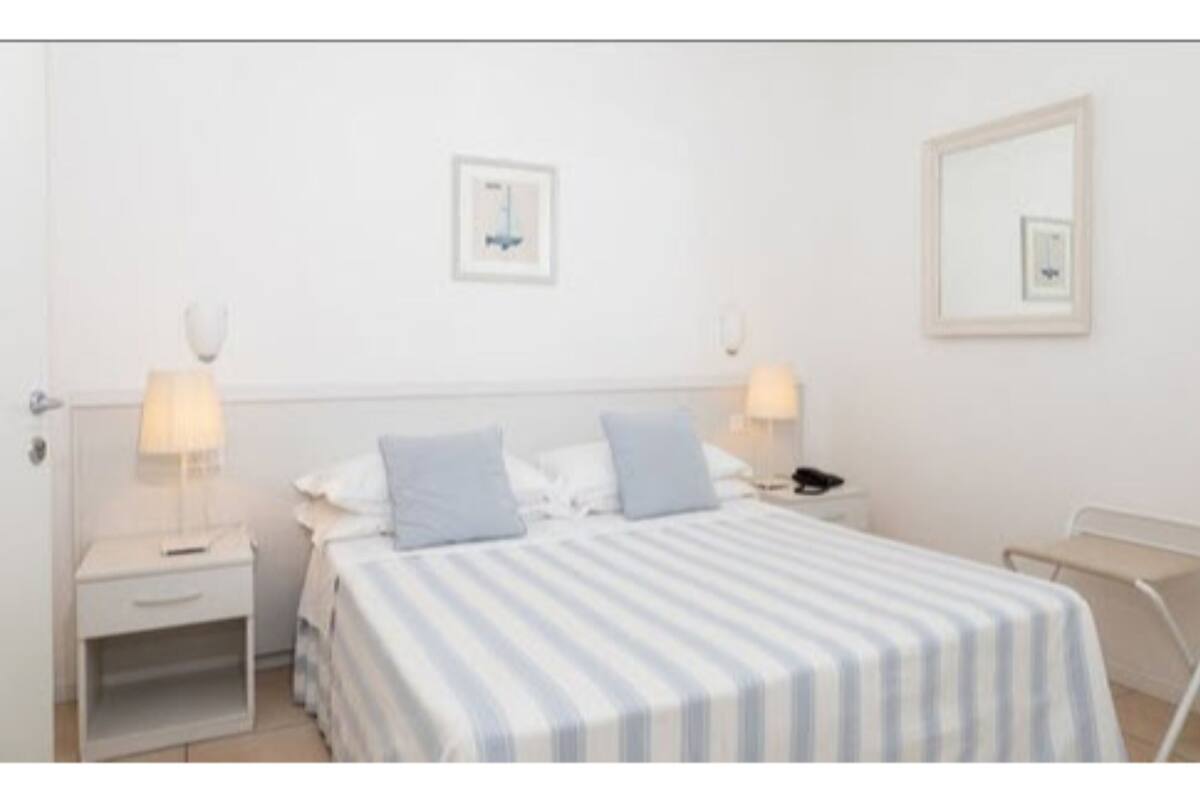 A hotel room with a bed, two bedside tables, a nightstand, a lamp, a mirror, and a small picture on the wall.