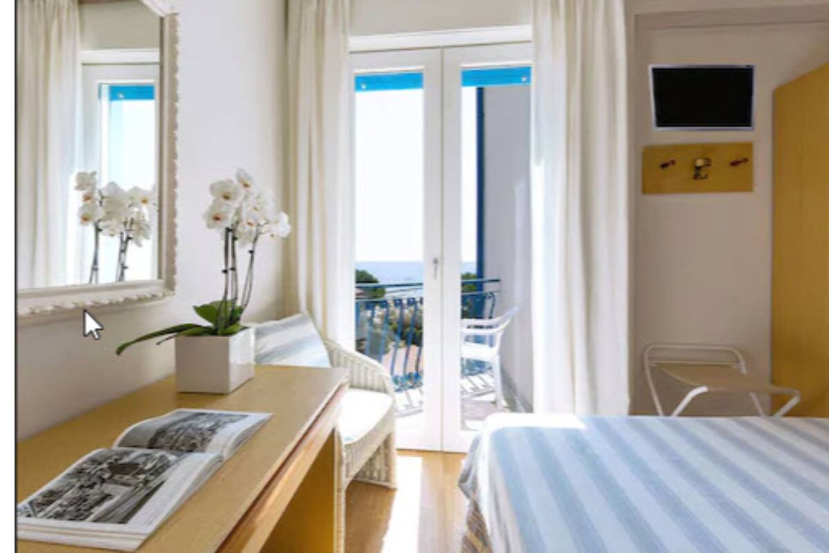 A hotel room with a bed, a desk with a book, a mirror, a window with curtains, a balcony with a chair, and a TV mounted on the wall.