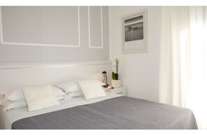 A neatly made bed with white linens and a gray blanket, a bedside table with a lamp and a potted plant, and a framed picture on the wall. - Hotel Plaza Esplanade (Jesolo)