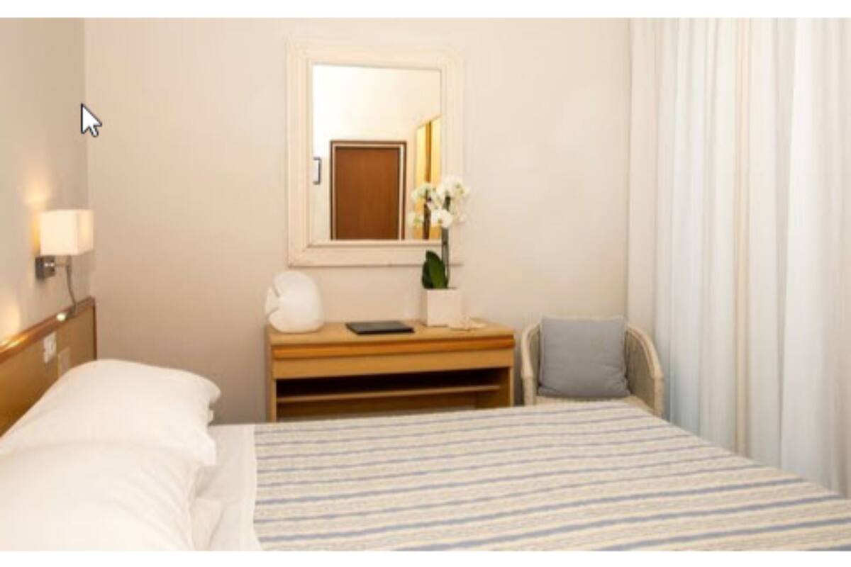 A hotel room with a bed, a bedside table with a lamp, a mirror, and a small desk with a chair.