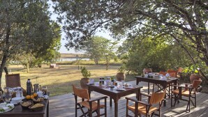 Un área de comedor al aire libre con mesas dispuestas para una comida, rodeada de árboles y con vista a un lago.