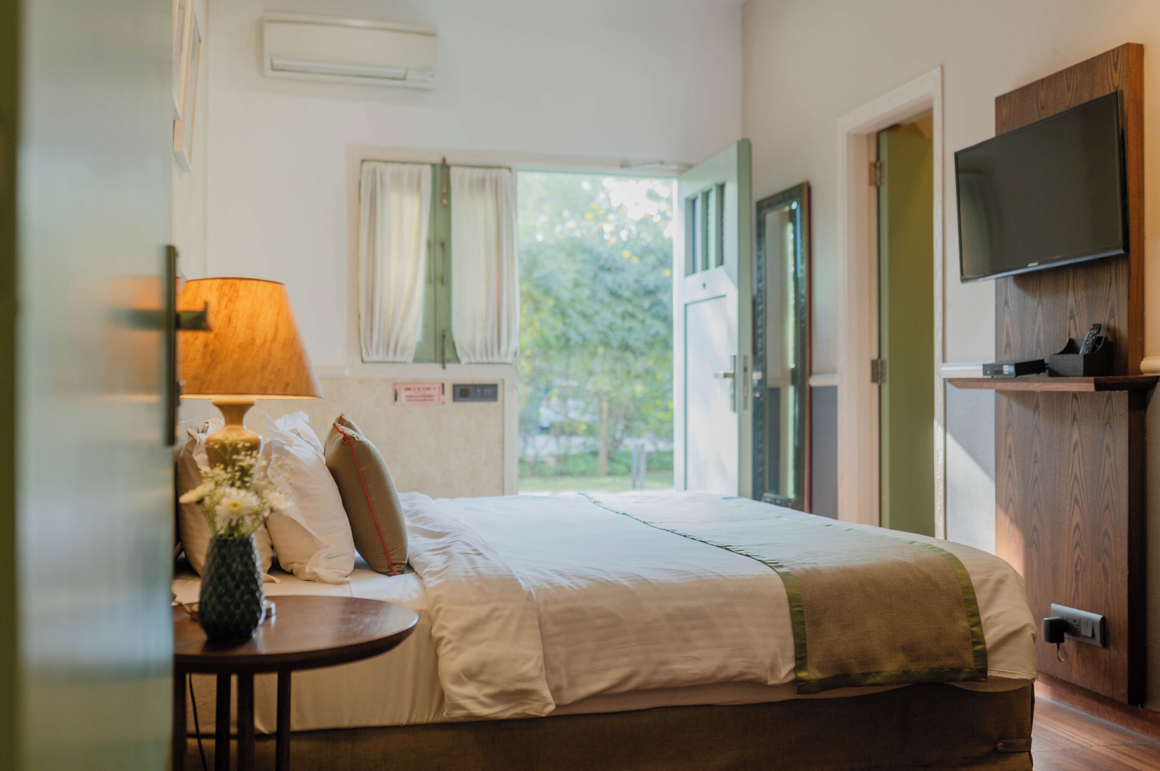 Habitación de hotel con cama, mesita de noche con lámpara, televisor montado en la pared y ventana con cortinas.