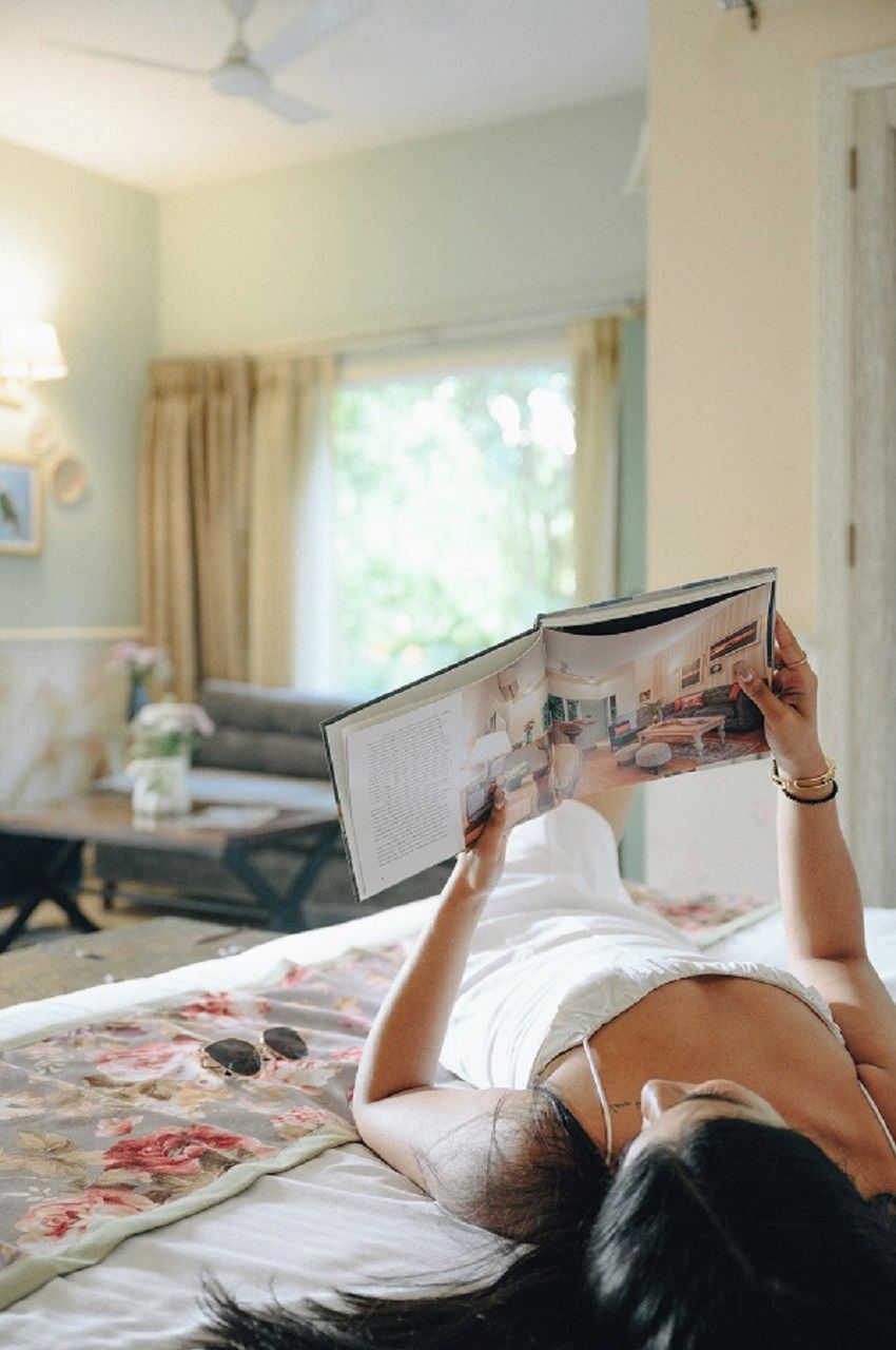 A person lying on a bed in a room with a ceiling fan, a window with curtains, and a lamp on a side table.