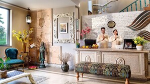 A well-furnished living room with a large window, a wooden accent wall, a decorative bench, and a reception desk with a decorative backdrop. - Karma Chalets (Gurugram)