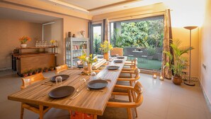 A dining area with a long wooden table set for six, surrounded by chairs. There is a bookshelf with various items on the left, and a large window with a view of greenery outside. - Karma Chalets (Gurugram)