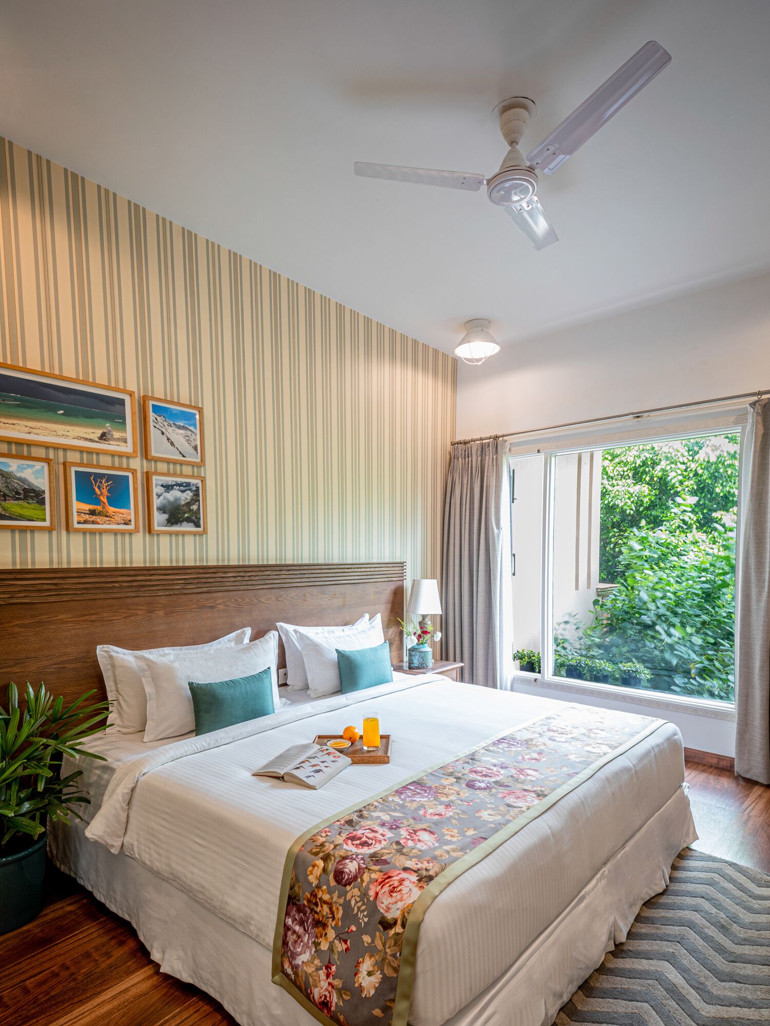 Una habitación de hotel con una cama grande, un ventilador de techo, una ventana con cortinas y cuadros en la pared.
