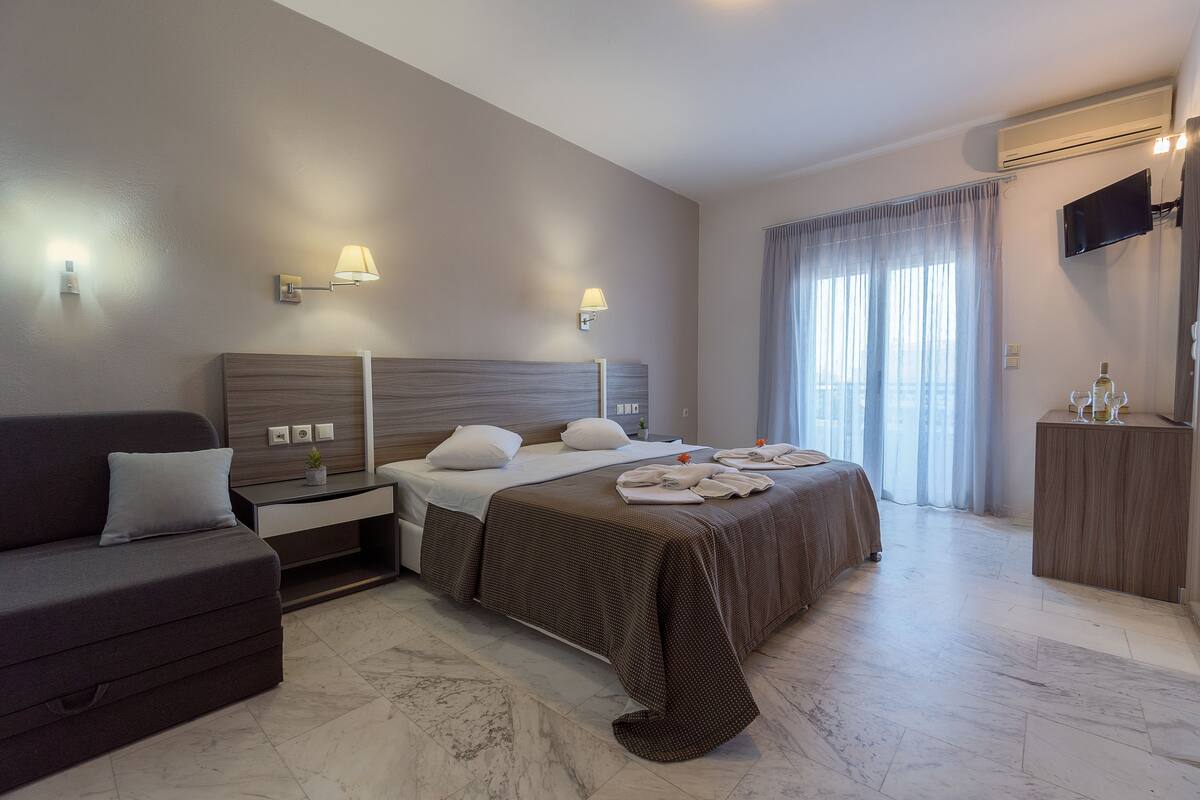A hotel room with a bed, a sofa, a TV, and a minibar.