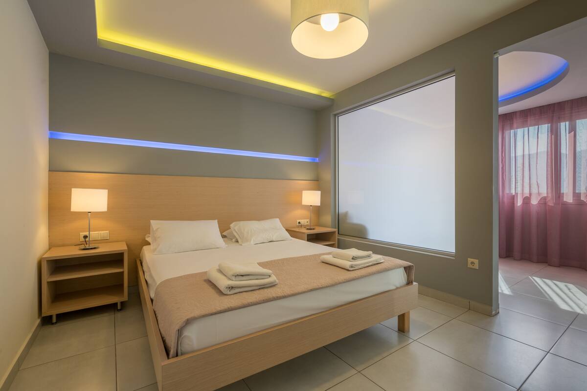 A modern hotel room with a large bed, bedside lamps, a nightstand, and a view of the outside through a large window.