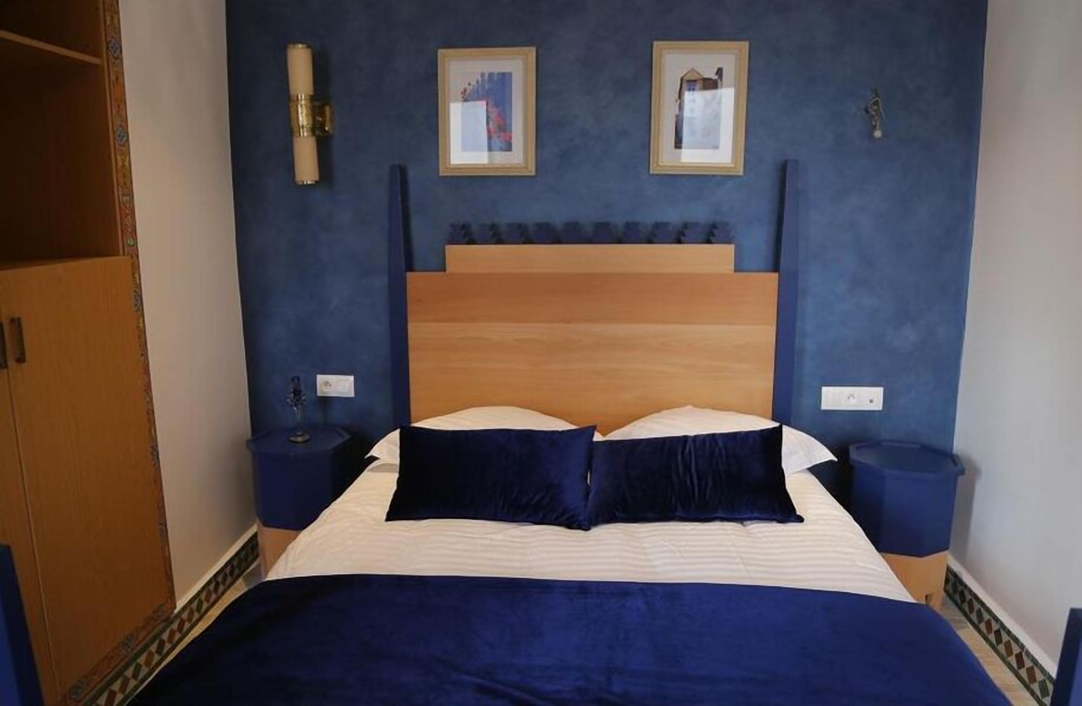 Un dormitorio con cabecera de madera, cama con ropa de cama azul oscuro, dos mesitas de noche y un armario.