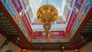 A grand hall with intricate red and gold patterns on the walls and ceiling, a large ornate chandelier, and a high ceiling with a skylight. - Palais Zahia (Tangier)