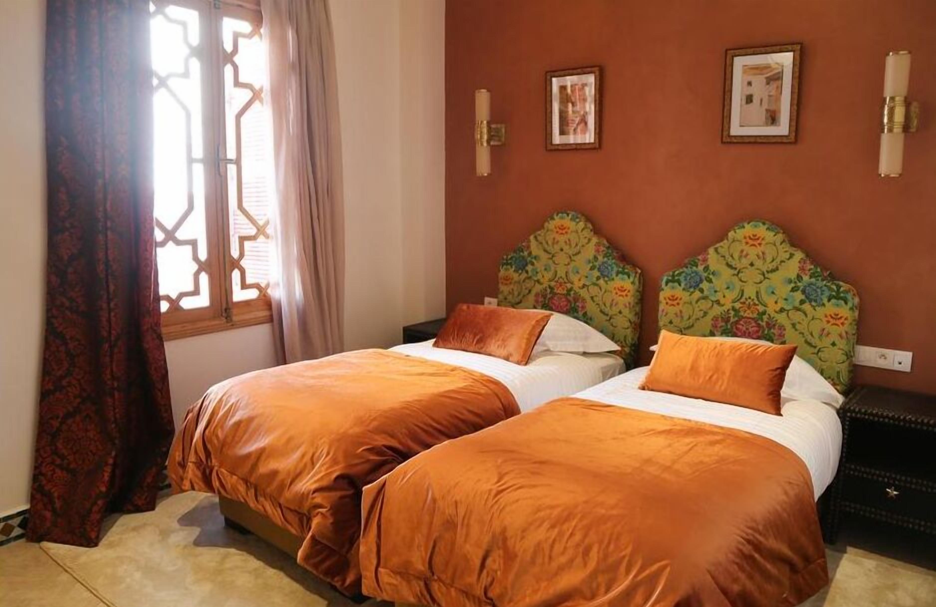 A room with two beds, each with matching orange bedding, a patterned headboard, and a window with a geometric design.