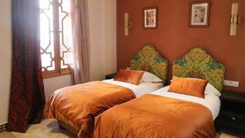 Una habitación con dos camas, cada una con ropa de cama naranja a juego, un cabecero estampado y una ventana con diseño geométrico.
