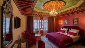 A room with a large bed, a chandelier, a red chair, a small table, and a mirror. - Palais Zahia (Tangier)