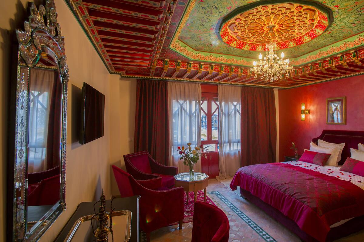 A room with a large bed, a chandelier, a red chair, a small table, and a mirror.