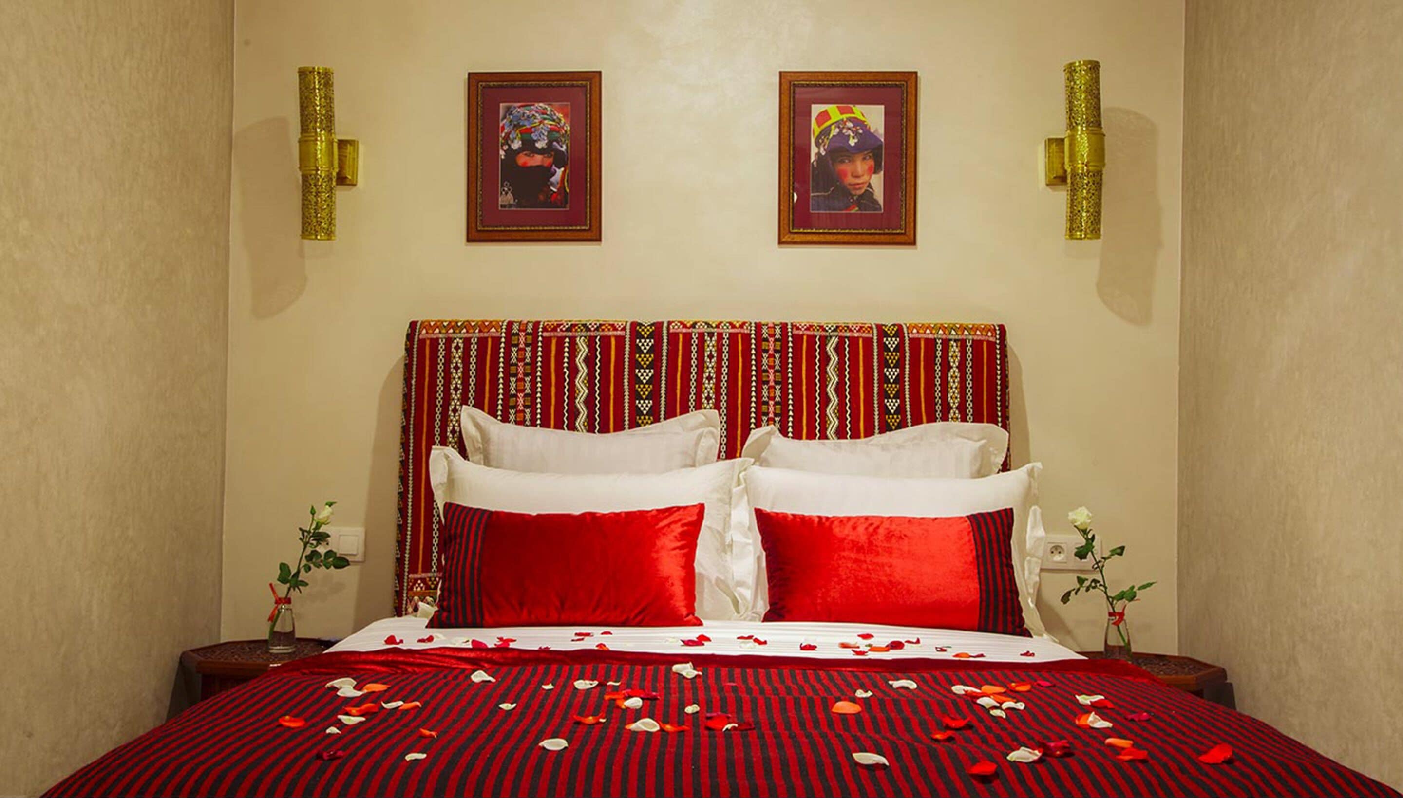 Una cama con ropa de cama roja y blanca, dos cuadros en la pared y apliques decorativos.