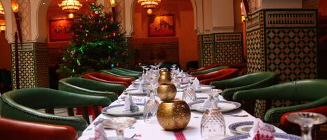 Un comedor con una mesa larga dispuesta para una comida, sillas tapizadas de verde y un árbol de Navidad al fondo.
