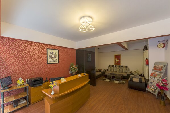 A reception area with a wooden desk, a computer, a printer, and a vase with flowers. There is a sofa and a coffee table in the background.