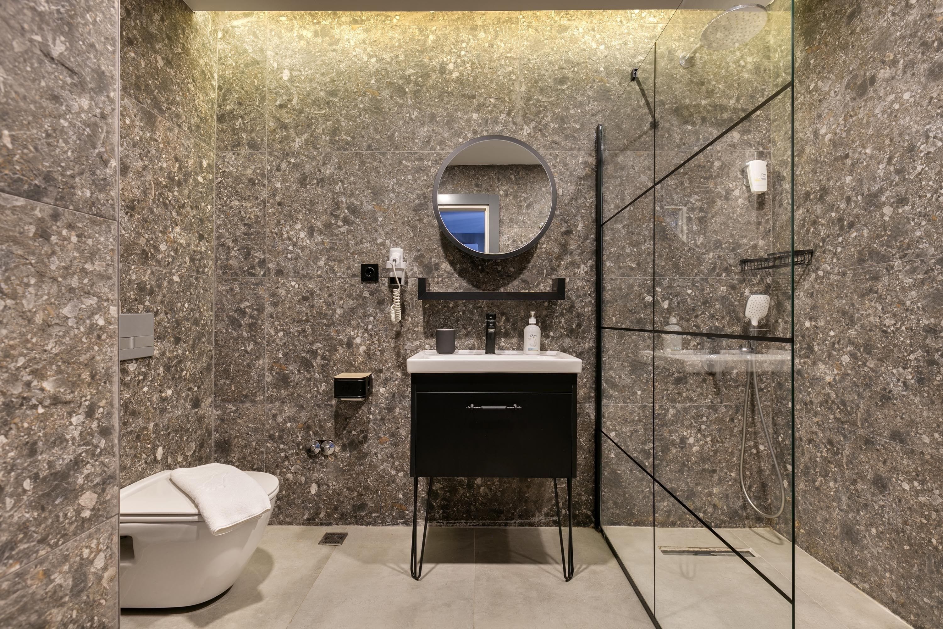 Ein modernes Badezimmer mit Marmorwand, einem runden Spiegel, einer schwarzen Waschtischkommode, einer Duschkabine aus Glas und einer weißen Toilette.