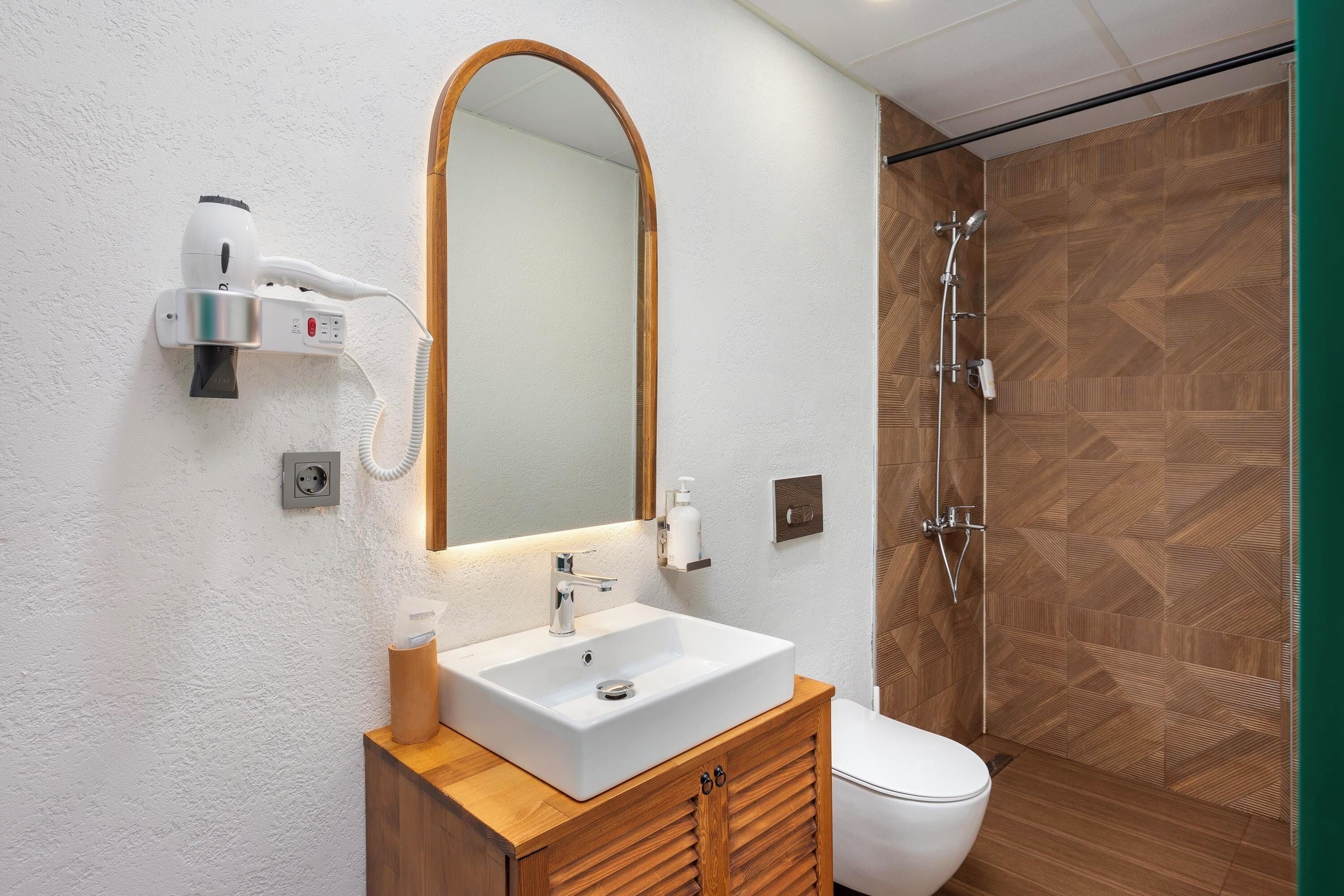 A bathroom with a white sink, wooden vanity, a large mirror, a hairdryer, and a shower with wooden walls.
