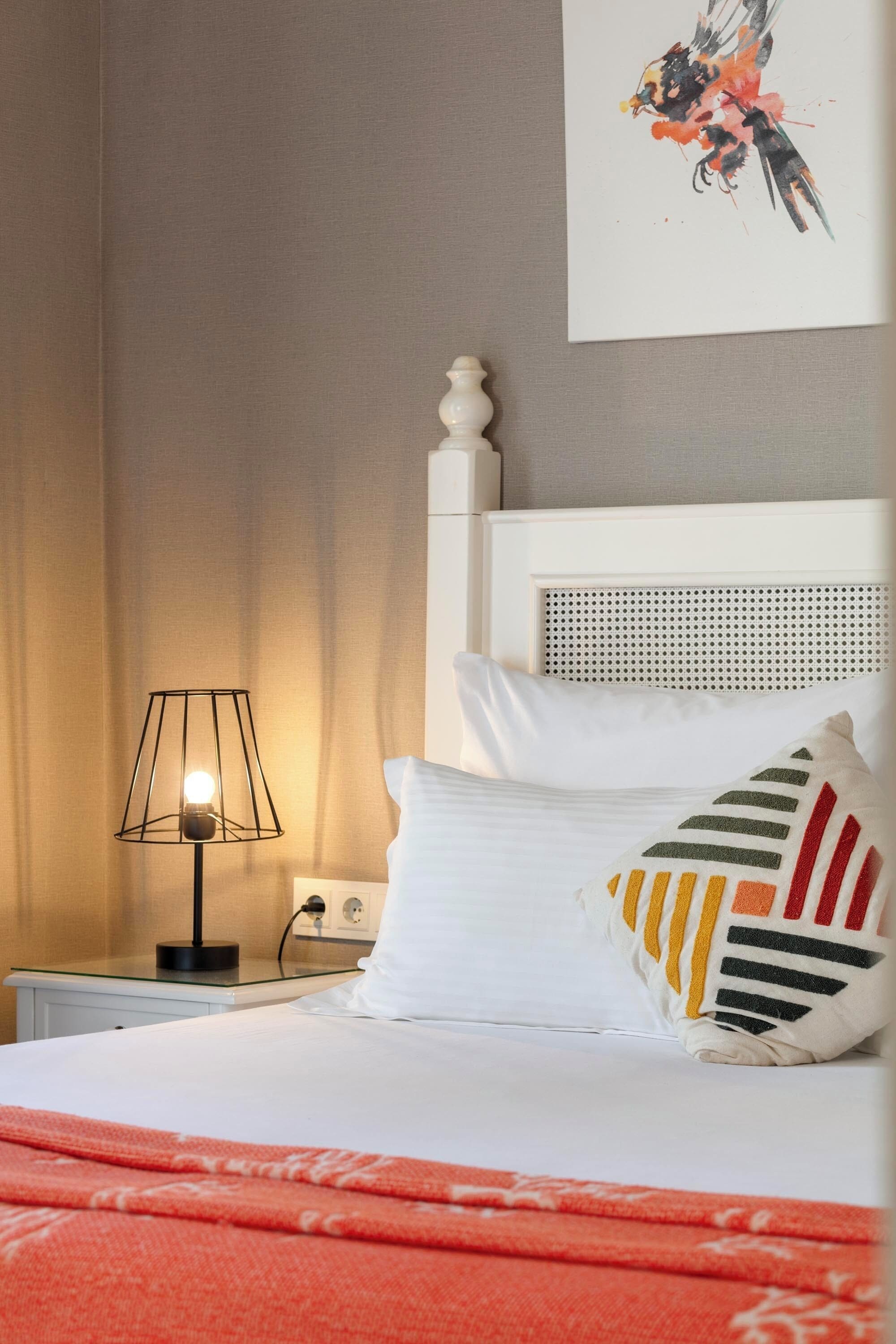 A neatly made bed with white linens, a striped pillow, and a red blanket. There is a bedside lamp with a black shade and a power outlet on the nightstand. A painting hangs on the wall above the bed.