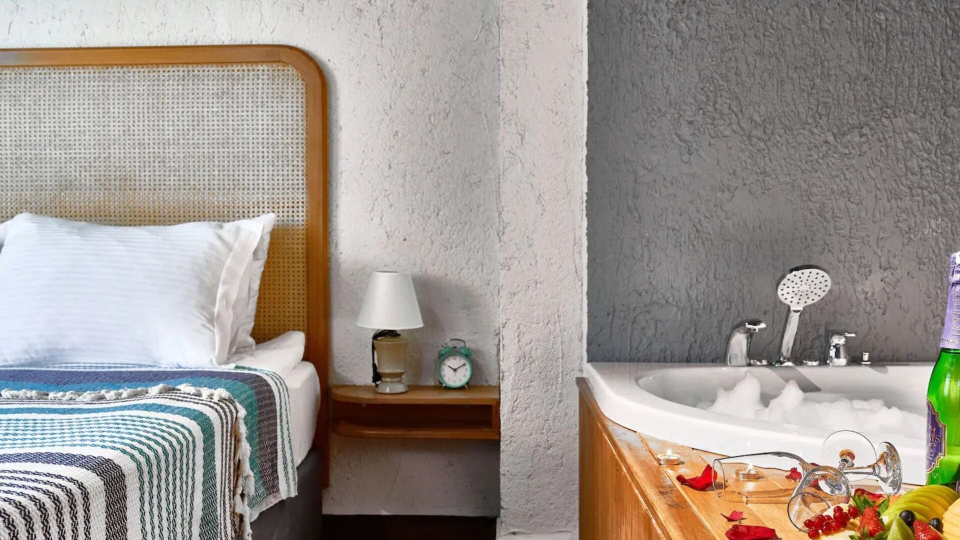 Habitación de hotel con cama, mesita de noche con lámpara, despertador y baño con bañera llena de burbujas.