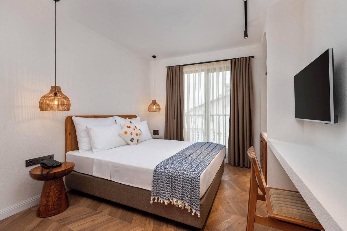A modern hotel room with a bed, bedside table, TV, and a window with curtains.