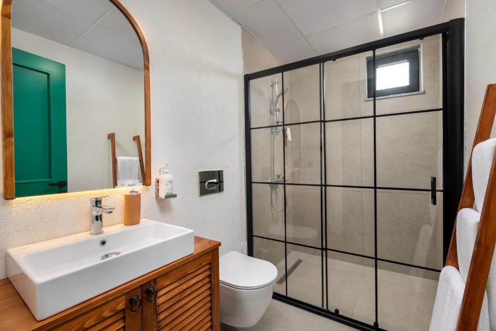Un baño con lavamanos blanco, tocador de madera, inodoro y zona de ducha con mampara de vidrio.
