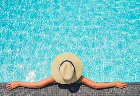 Una persona recostada al borde de la piscina con los brazos extendidos, usando un sombrero, y una piscina con agua cristalina azul.