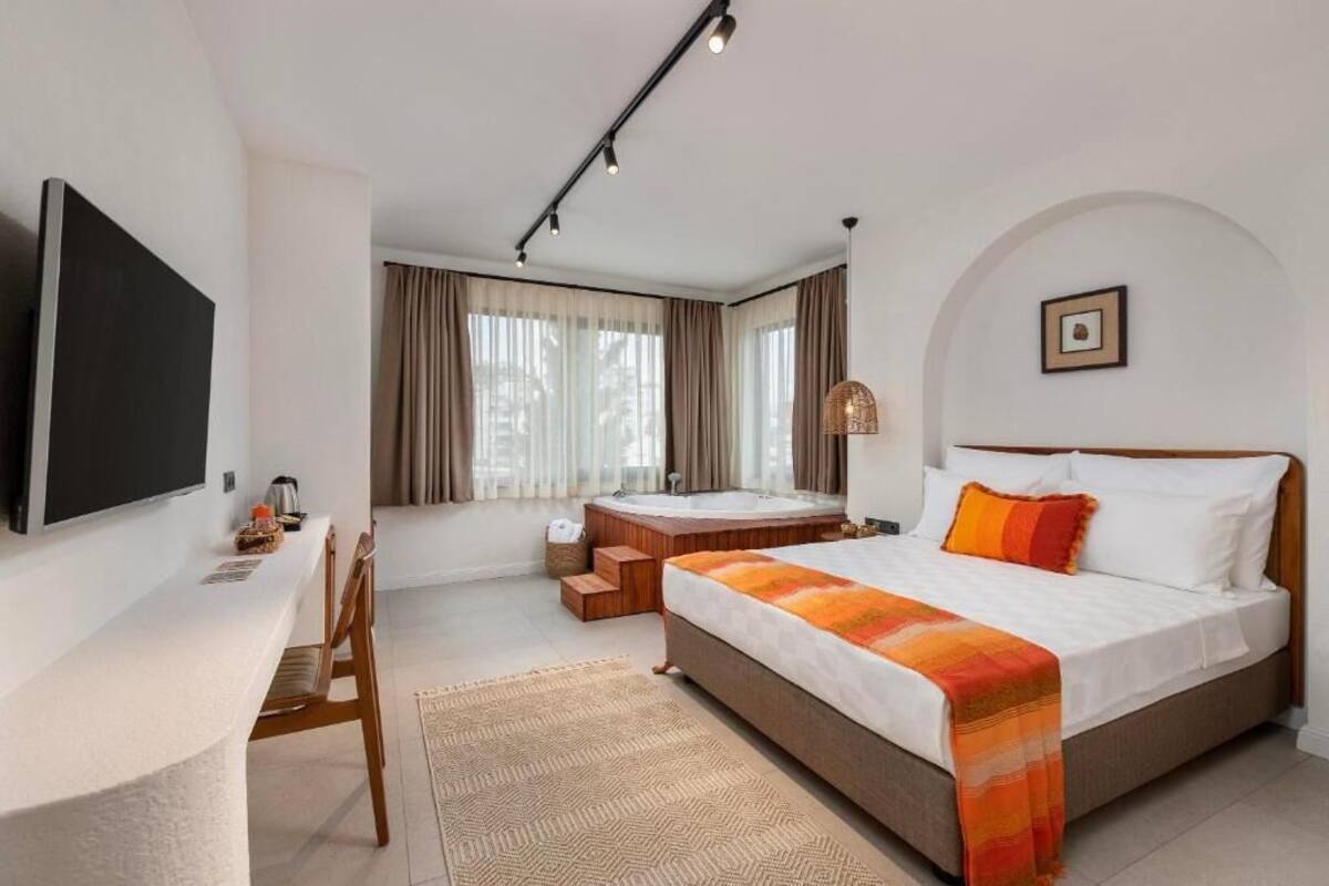 A modern hotel room with a large bed, a desk with a flat-screen TV, a bathtub, and a large window with curtains.