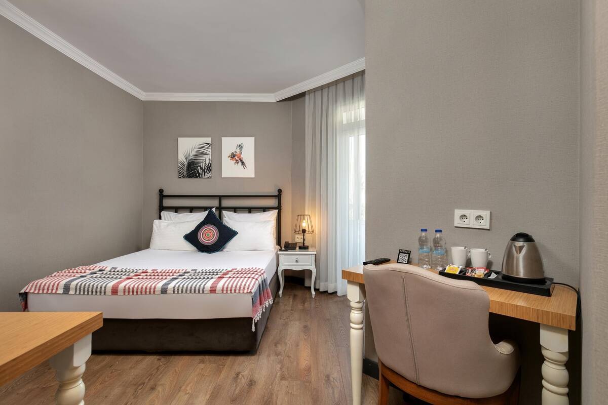 A hotel room with a bed, bedside table, desk, chair, and artwork on the wall.
