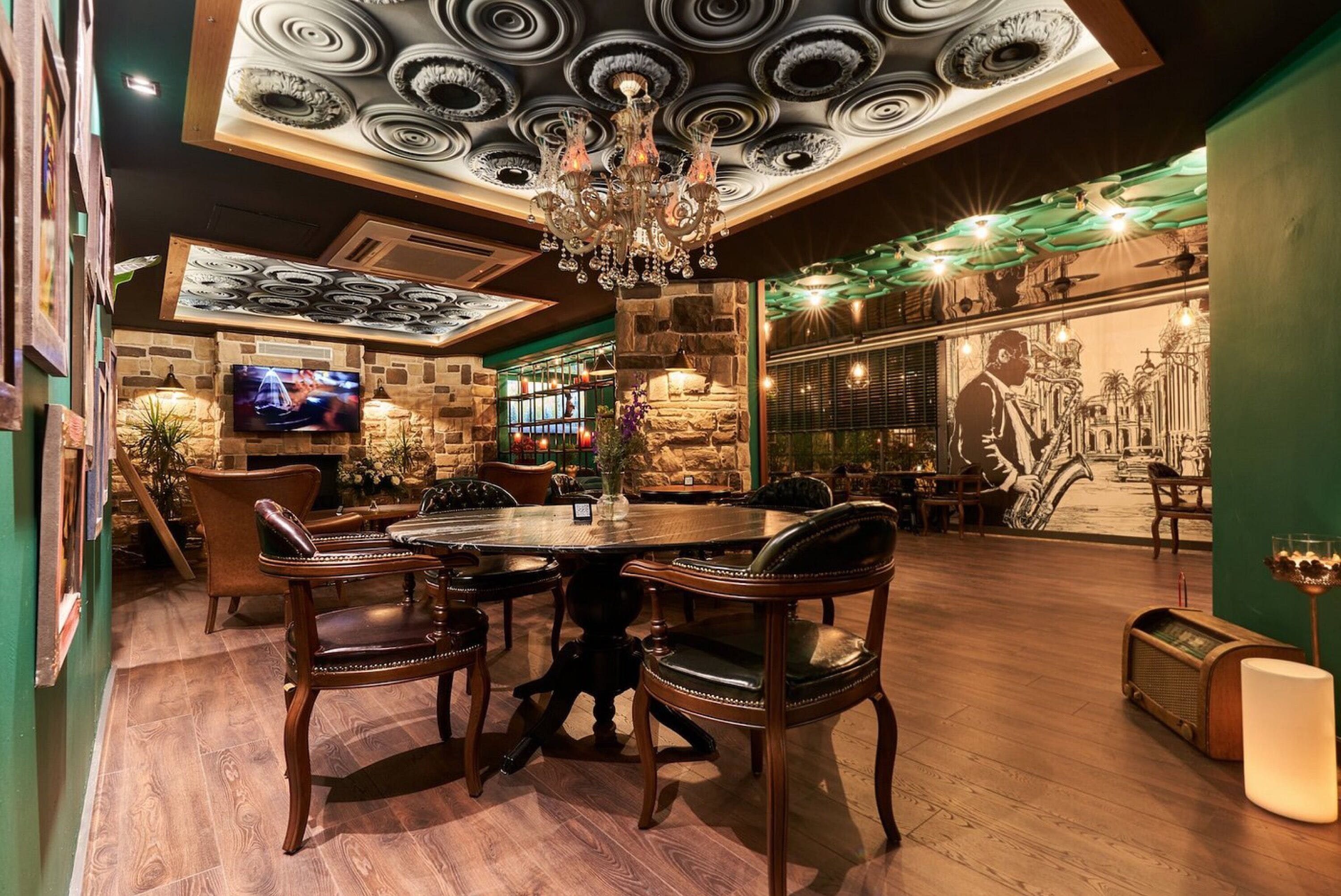 A restaurant with a round table, wooden chairs, a chandelier, a television, and a mural on the wall.