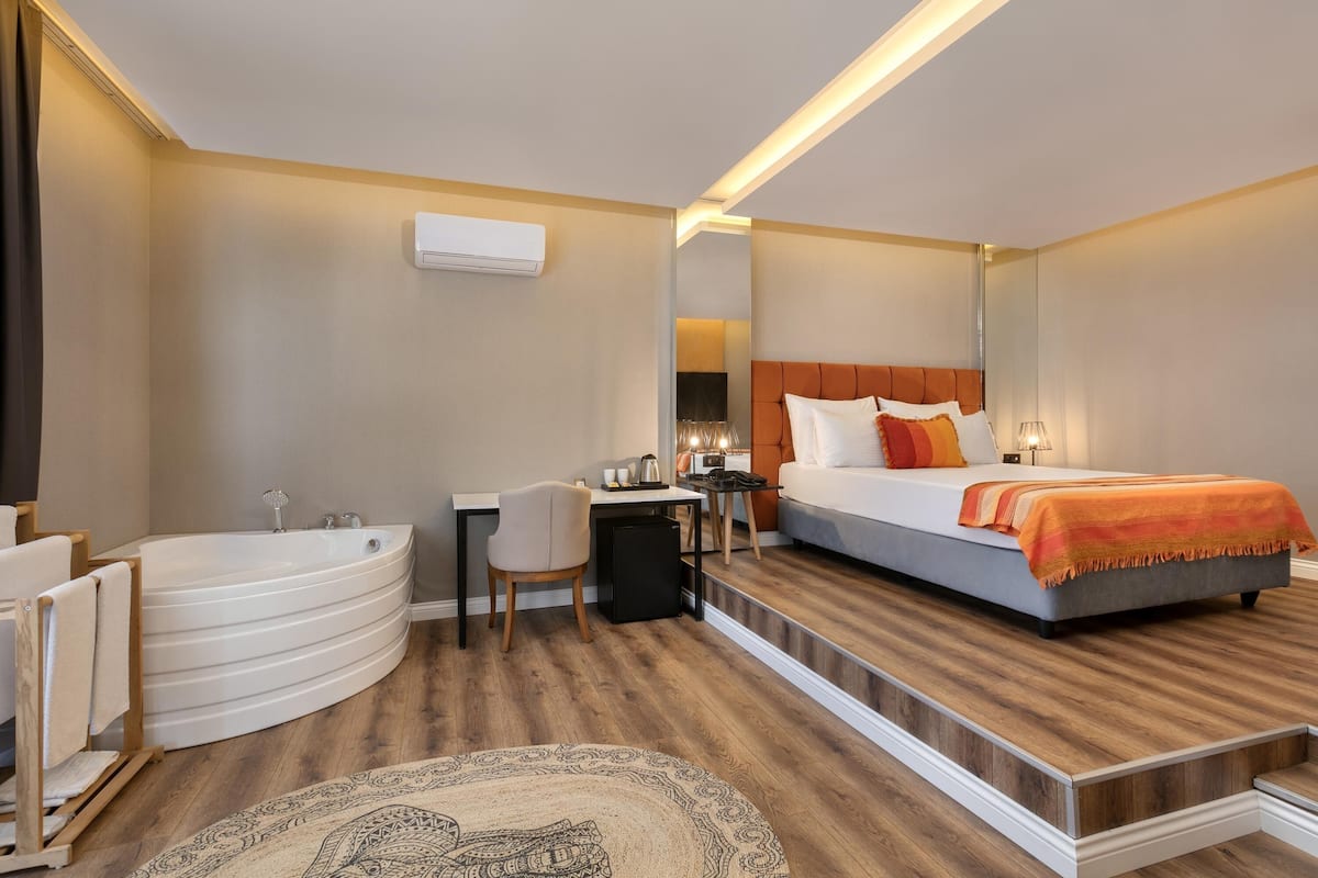 A modern hotel room with a curved bathtub, a desk with a chair, a bed with an orange blanket, and a large mirror.