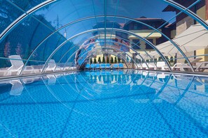 Piscina azul límpida com teto de vidro, espreguiçadeiras e vista para edifícios ao fundo.