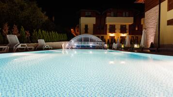 Une piscine entourée de bâtiments dont les fenêtres sont illuminées, avec des chaises longues et des parasols à proximité.