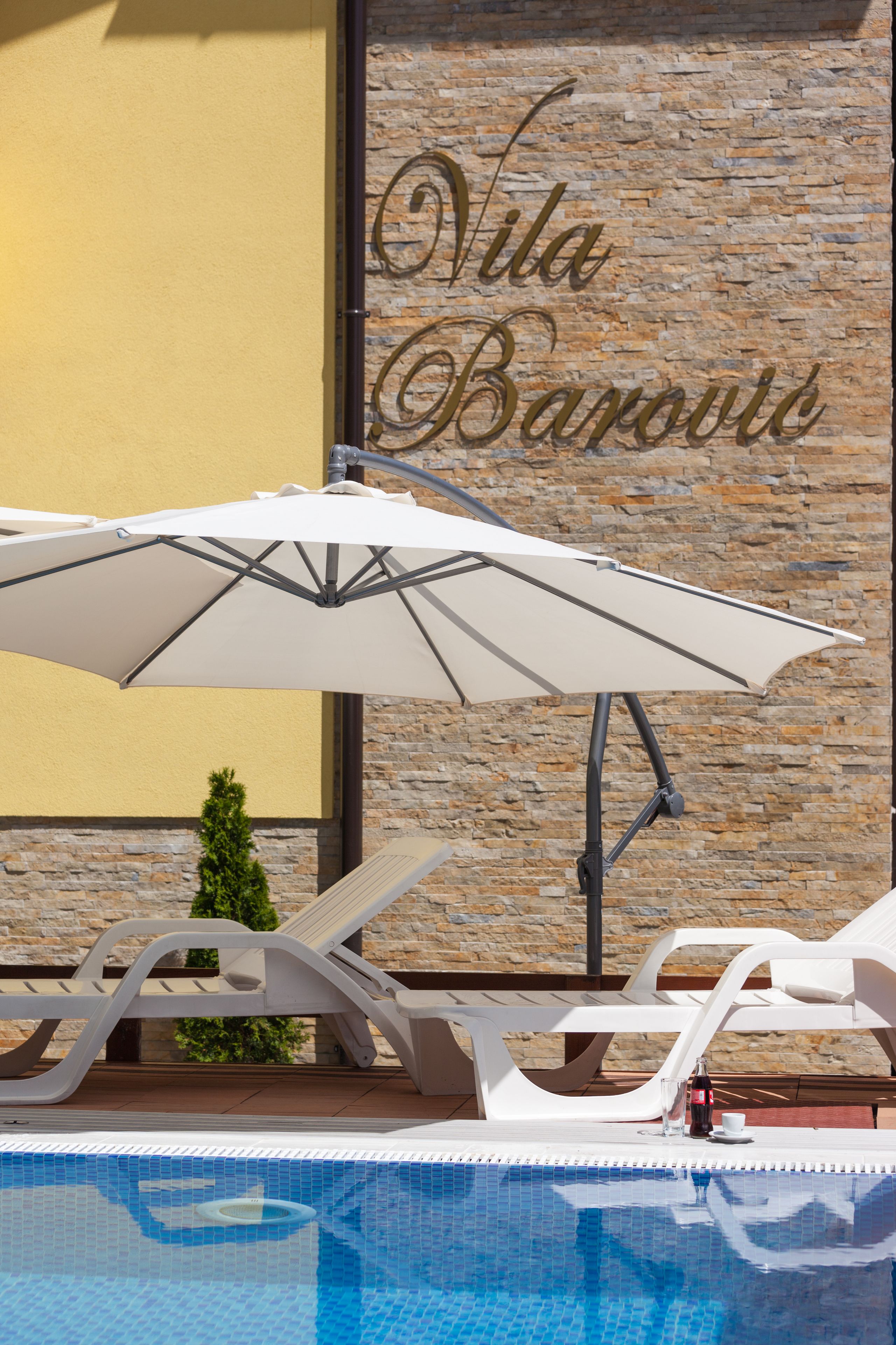 A pool area with lounge chairs and an umbrella, a stone wall with the text "Vila Barouce", and a small potted plant.