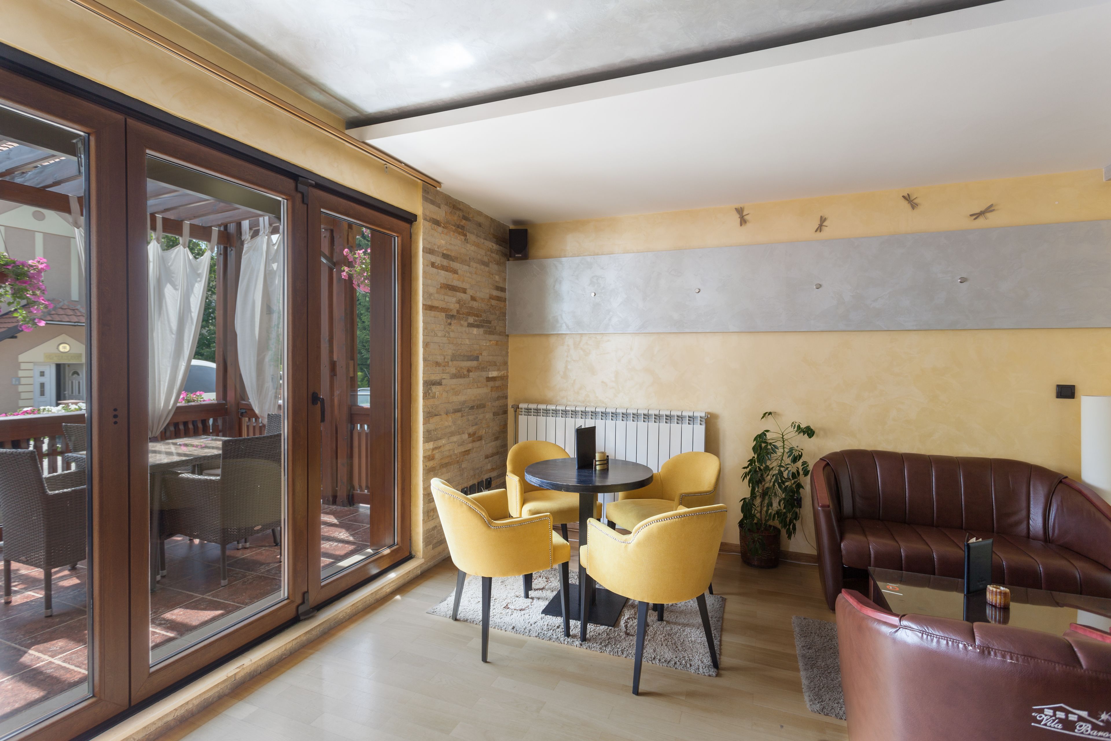 Een moderne woonkamer met een glazen deur die leidt naar een terras, een ronde salontafel, gele stoelen, een bruine leren bank en een potplant.