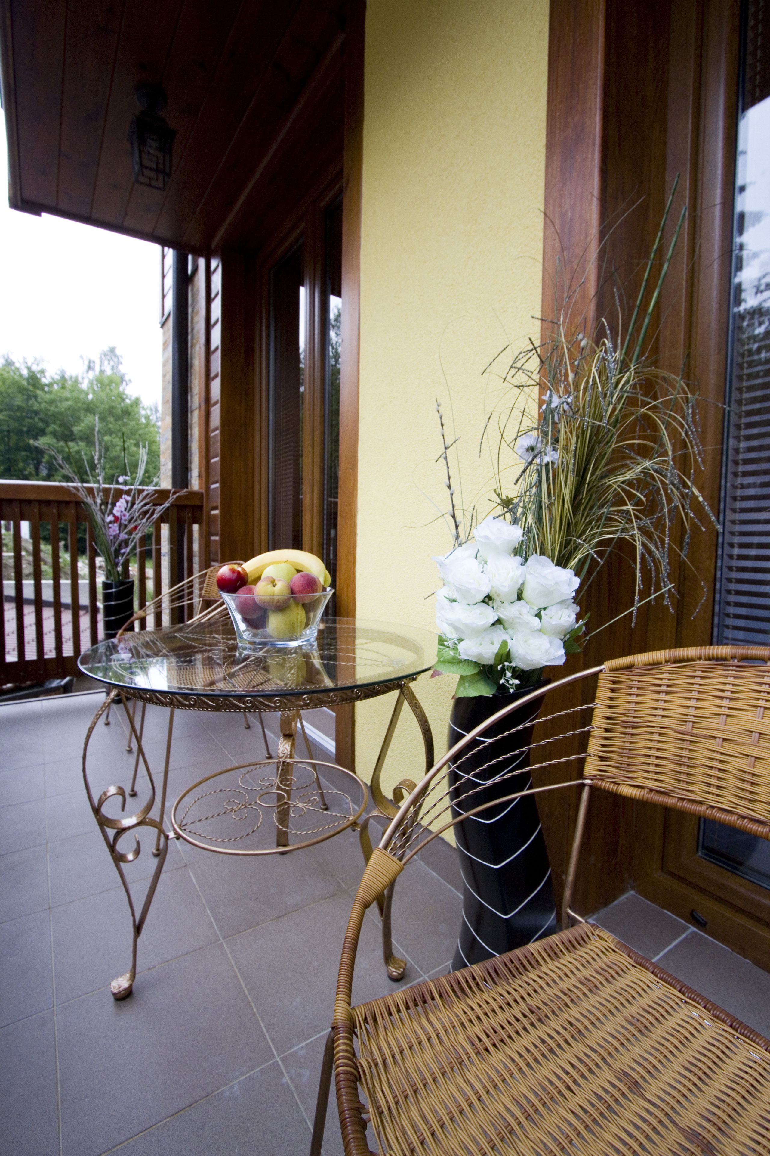 A glass table with a bowl of fruit, a wicker chair, and a vase of flowers on a balcony.