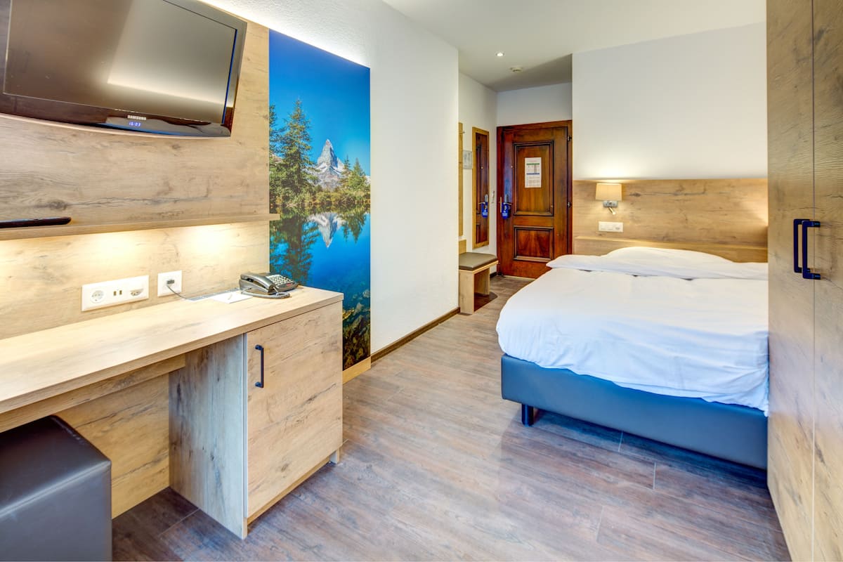 A hotel room with a wooden desk, a bed, a TV, and a mural of a mountain.