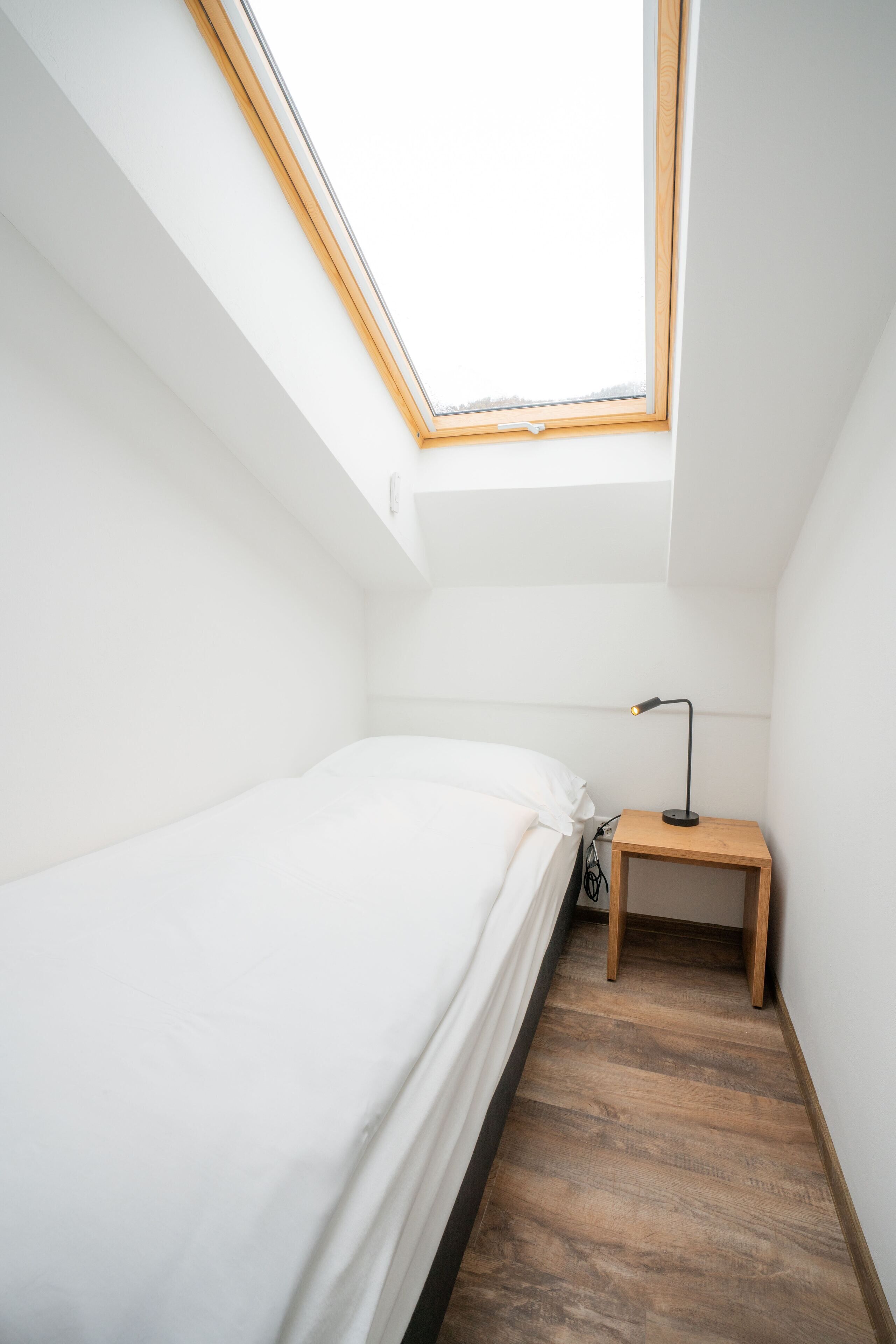 A small, bright room with a skylight, a single bed, a bedside table with a lamp, and a wooden floor.