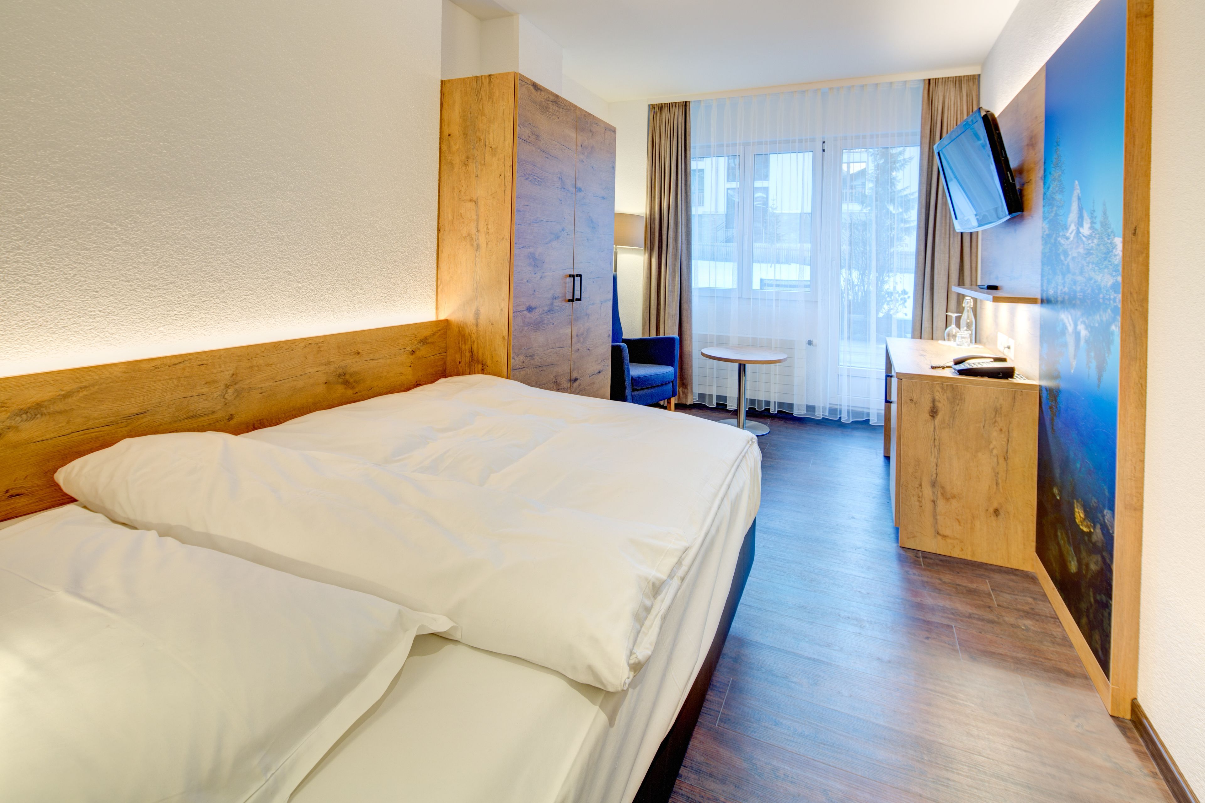 Une chambre d’hôtel avec un grand lit, un bureau avec une chaise, un téléviseur fixé au mur et une fenêtre avec des rideaux.