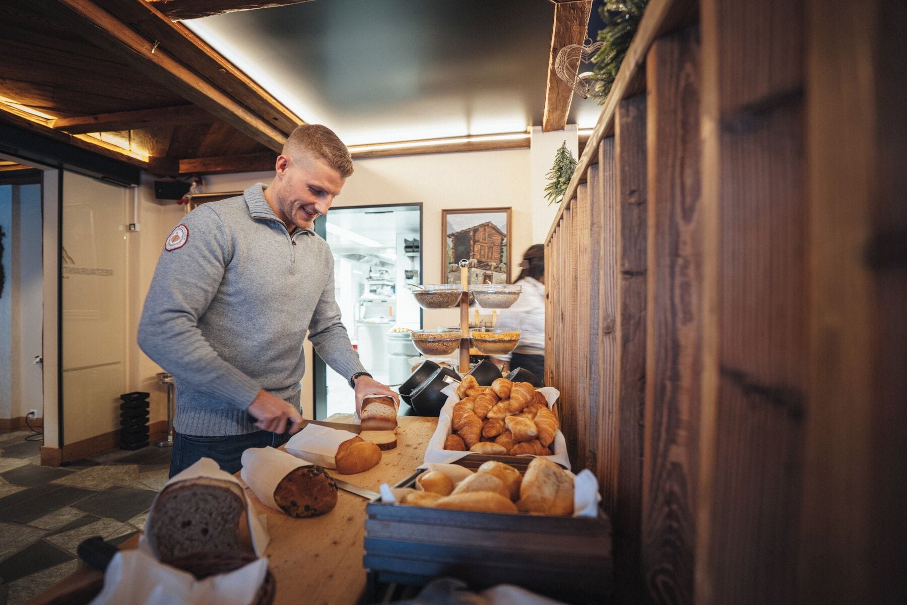 Una persona cortando pan en una panadería con interiores de madera y una exhibición de pasteles.