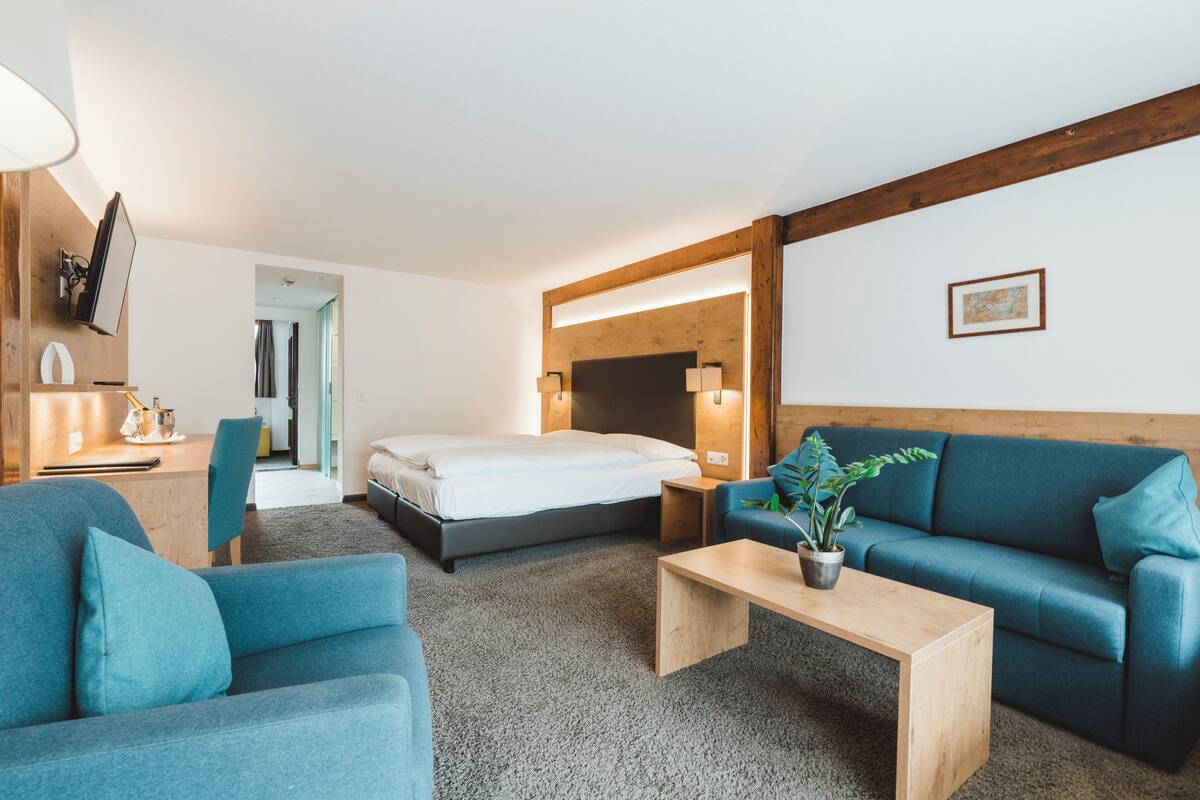 A modern hotel room with a bed, a blue sofa, a wooden coffee table, and a desk with a lamp.