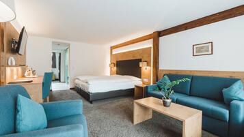 A modern hotel room with a bed, a blue sofa, a wooden coffee table, and a desk with a lamp.