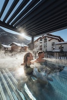 Una pareja en un jacuzzi con vapor ascendente, rodeada por una terraza de madera y un edificio con techo inclinado en un paisaje nevado.